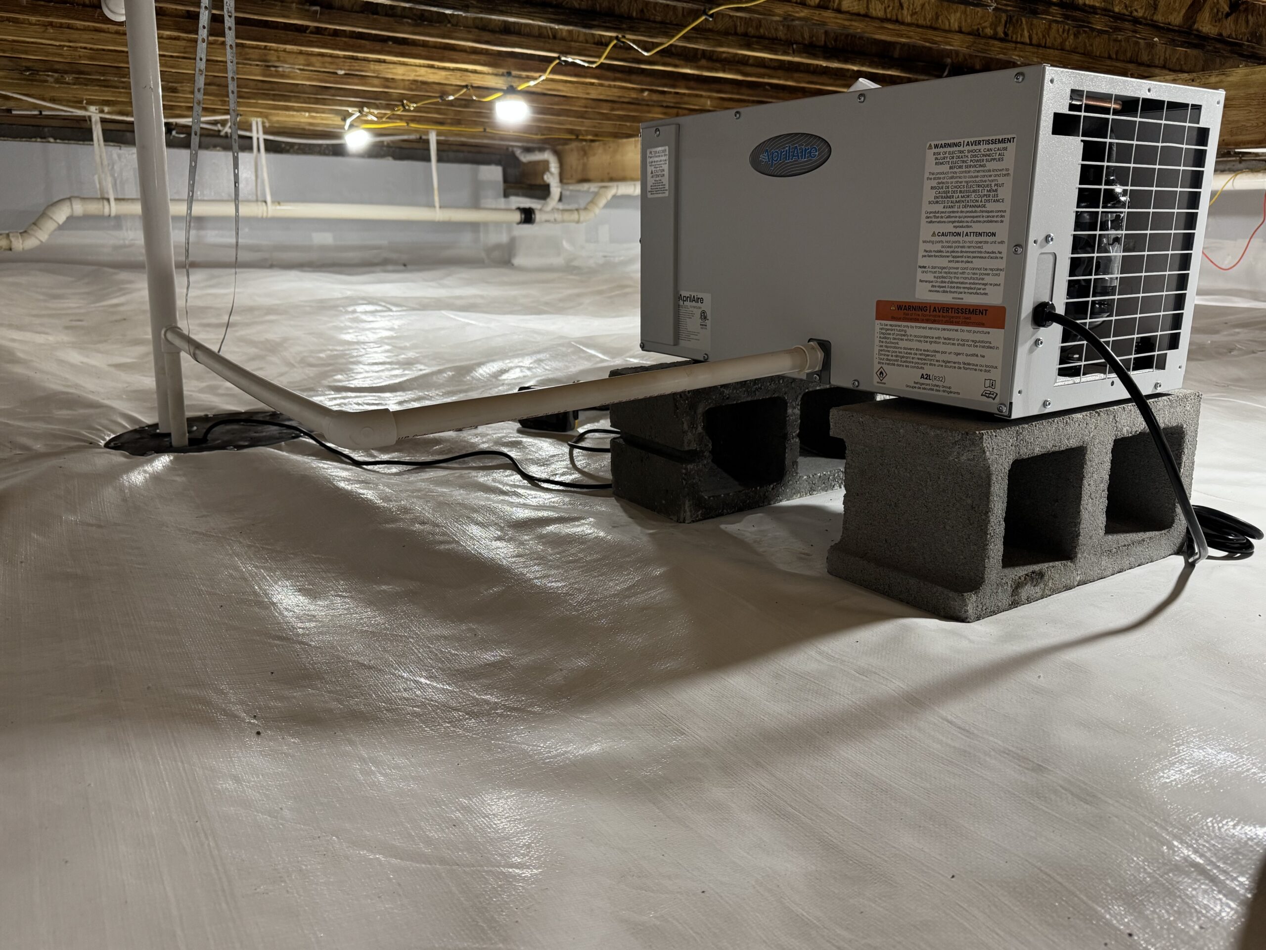 A dehumidifier sits on cinder blocks in a plastic-lined crawl space with exposed wooden beams and overhead lighting.